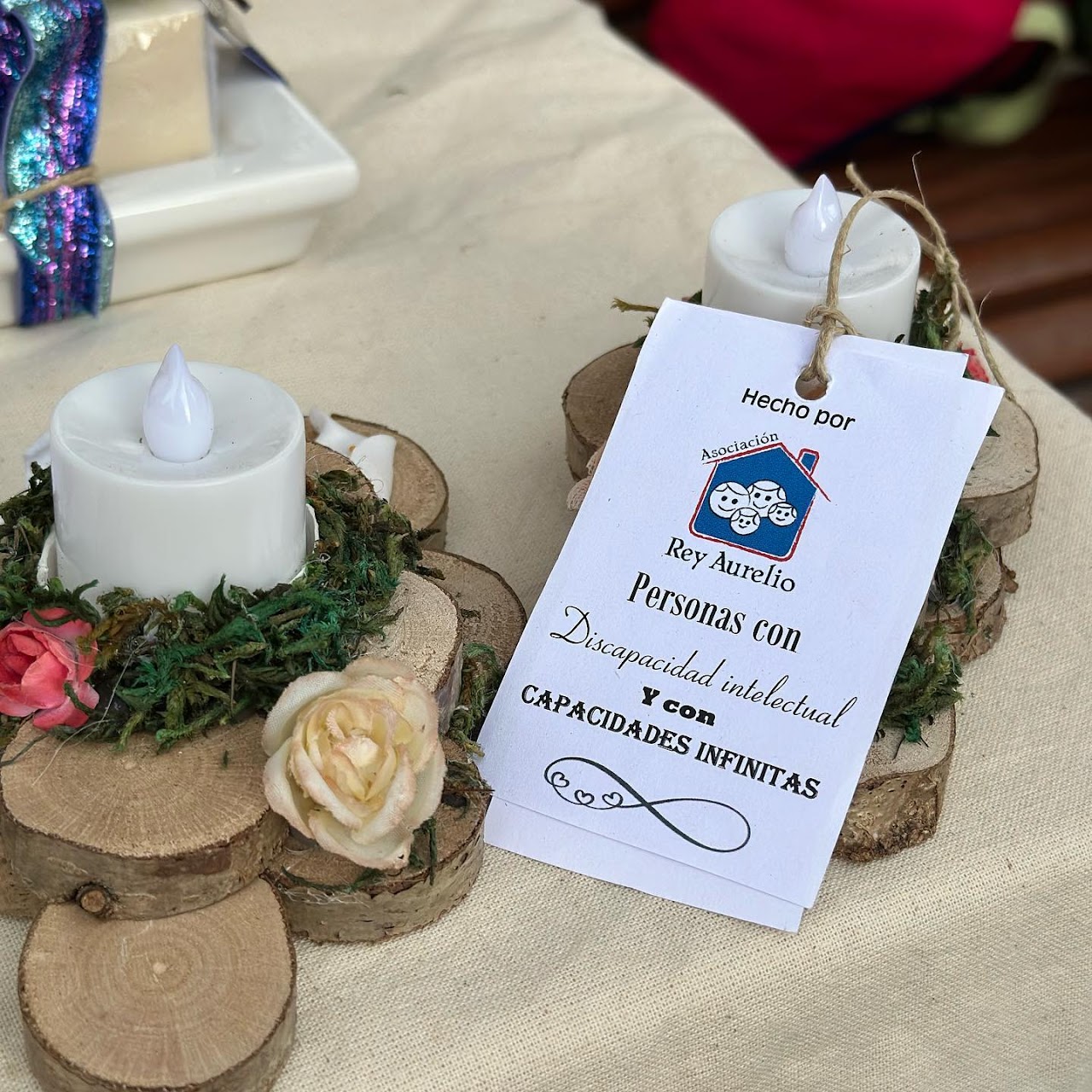 Velas decoradas sobre rodajas de tronco con musgo y flores, con una etiqueta «Hecho por Asociación Rey Aurelio. Personas con discapacidad intelectual y con capacidades infinitas»